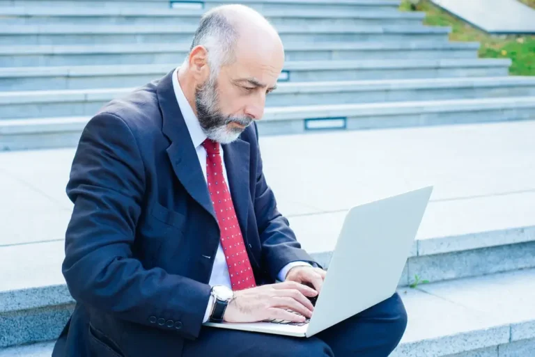 Thoughtful businessman using laptop while sitting on stairs. Focused middle aged attorney typing on laptop outdoors. Technology, business concept