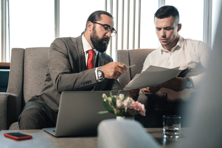 Giving pen. Bearded businessman giving pen to his partner signing contract sitting on the sofa