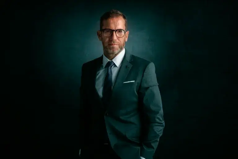 Portrait of wrinkled face businessman wearing suit and eyeglasses while standing against at isolated dark backgorund.